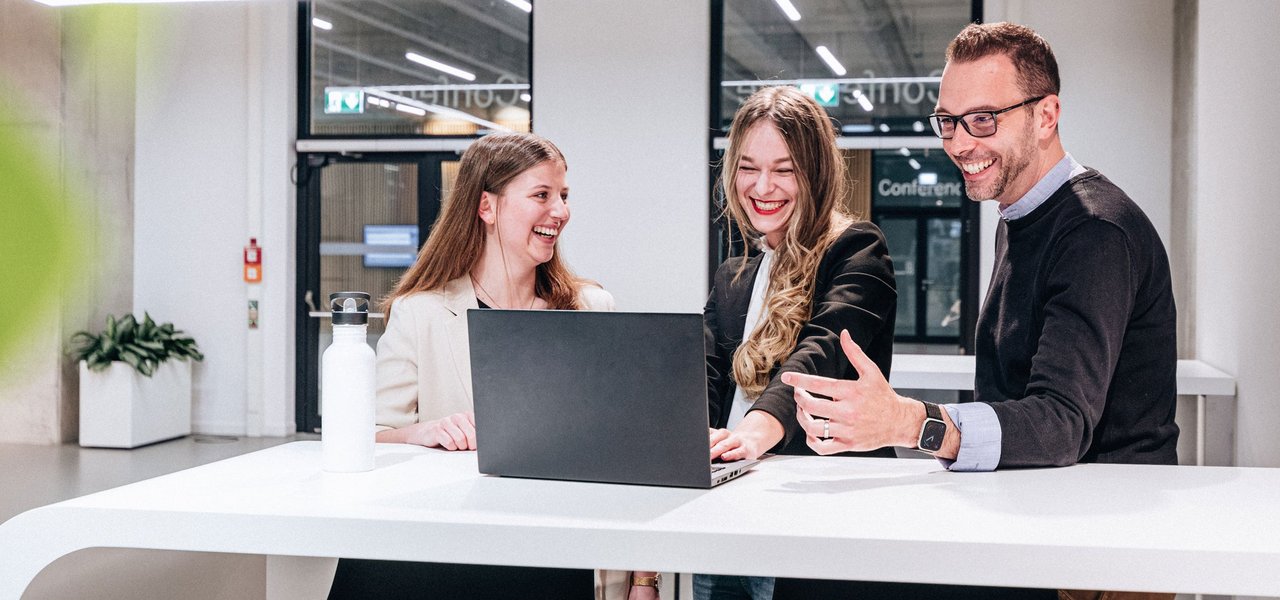 Drei Mitarbeitende lachen gemeinsam bei einer Besprechung an einem modernen Arbeitsplatz. Das Bild steht für gelebte Teamkultur und die attraktiven Benefits, die bei Daimler Truck zu einem motivierenden Arbeitsumfeld beitragen.