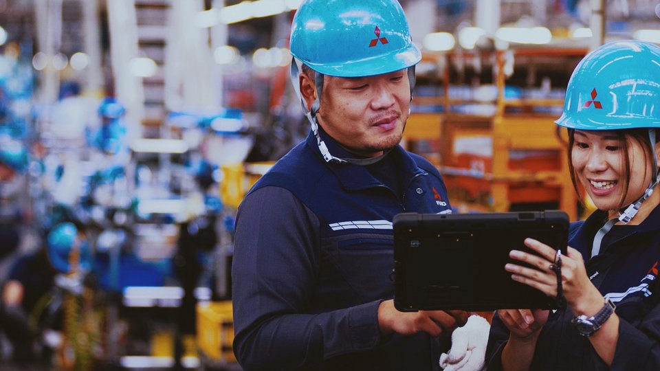 Two production employees wearing blue hard hats look at a process monitoring tablet in a busy production hall.