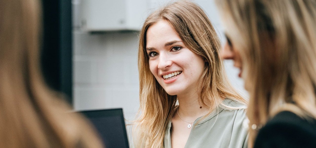 A young woman smiles and looks round as she sits with colleagues at a table with a laptop.